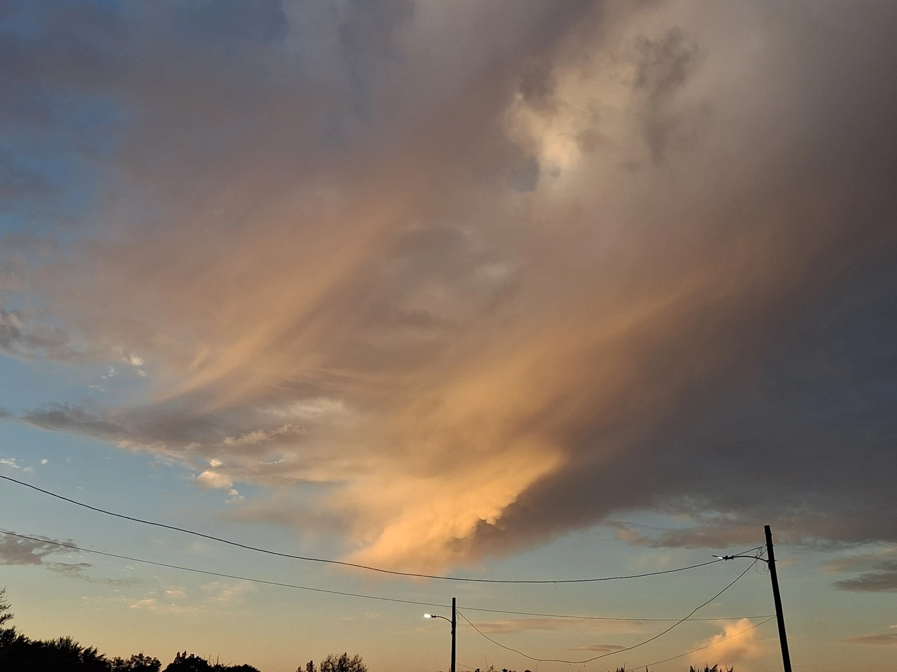 A sweeping golden cloud formation above a treeline and overhead utility lines.